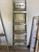 Front view of the metal step ladder leaning against the wall, showing five steps and the top platform with circular holes.