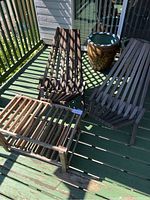 Two wooden deck chairs and one wooden slatted side table arranged on a green painted wooden deck with railing, showing natural wood finish and slatted design.