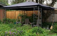 Gazebo set up in backyard with grey soft roof and metal frame pillars with leaf design. Two wooden chairs inside.