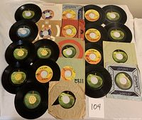 Photo showing a collection of 22 vinyl 45 RPM records with various sleeves and labels including Apple and Capitol Records. Records arranged on a white background.