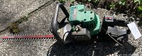 Full side view of the 22-inch gas hedge trimmer showing green engine body, black handle, and red steel blade lying on concrete.