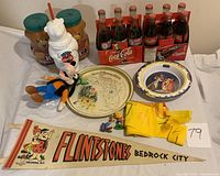 Full view of lot items showing 1999 Pan-Am Games and Saskatchewan Centennial Coca-Cola 6-pack, plastic polar bear cup, Kraft peanut butter jars, Disney and Flintstones memorabilia, plush Goofy toy, and McDonald's bibs.