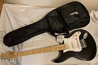 Full view of Behringer black strat style electric guitar lying next to black soft case on white fabric background.