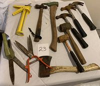 Full view of tools including caulking dispenser, hatchet, pickaroon, hammers, mallets, and hedge clippers arranged on a white surface.