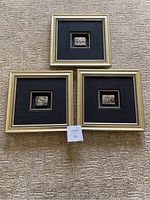 Three square gold-tone frames with black fabric mats and central silver-tone metal relief plaques on carpet