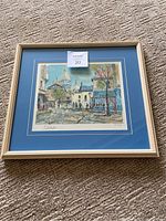 Framed Montmartre-Place du Tertre I painting by Eugene Pierre Cambier laying on carpet floor. Shows full view of frame and matte.