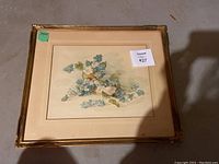 Top-down view of framed botanical print with gold frame and cream mat, displaying blue and white flowers.