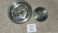 Both decorative pewter plates resting on carpet, front view showing detailed embossed scenes and ornate rims.