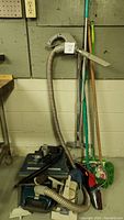 Photo showing Kenmore Whispertone vacuum with hose and wand, Dirt Raider vacuum, and floor cleaning tools grouped vertically against a wall.