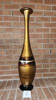 Tall glass decorative vase with gold and black finish against a brick wall background.
