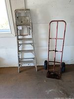 Full view of 5-foot aluminum ladder and handle dolly side by side against a white wall with a window and pegboard