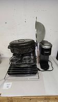 Photo showing metal mesh strainer, enamel oven roaster pan, broiler rack pan, muffin pans, grilling tops stacked, and Keurig K-Mini coffee maker