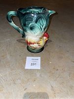 Photo of ceramic pitcher featuring a green and multicolor rooster design on one side, with handle and spout visible.