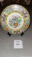 Front view of porcelain plate on black stand, showing detailed floral and bird pattern with yellow border.