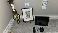 Weather station thermometer hanging on the wall, framed painting on wooden stand, and digital photo display on carpeted floor.