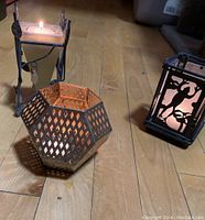 Photo showing three of the candleholders placed on a wooden floor, featuring a lit candle in a square metal holder with hanging charms, a hexagonal metal holder with a diamond cutout pattern, and a glass holder with silhouette cutout design.