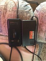 Photo showing two black rectangular 60V e-bike chargers placed side by side on a couch; one charger shows indicator lights labeled with battery percentages and the other shows a label with technical specifications.