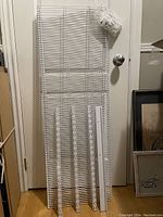 Two white wire closet shelves hung vertically on a door with four white vertical mounting runners leaned in front and a sealed bag of white plastic fasteners hanging at the top right.