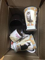 Eight coffee mugs in a box showing two cat motif mugs, one Starbucks France espresso cup and two coffee colored mugs.