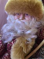 Close-up of Santa's face depicting detailed facial features, blue eyes, curly white beard, and golden brown faux fur hat.