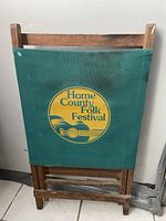 Front view of the folding wooden chair showing green fabric seat back with 'Home County Folk Festival' logo.