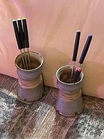 Six metal fondue skewers inserted into two ceramic holders. Each skewer has a black handle with a colored tip and the holders are cylindrical with a glazed ceramic finish.