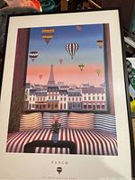 Full view of framed poster featuring Paris cityscape, Eiffel Tower and colorful hot-air balloons at dusk with striped cushions in foreground.
