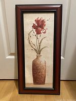 Front view of the framed wall art hung against a door, showing the vase with red flowers in a brown wooden frame.