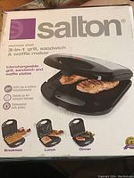 Photo of the product box showing the Salton 3-in-1 grill, sandwich, and waffle maker in use, grilling meat with the appliance closed, highlighting use cases for breakfast, lunch, and dinner.
