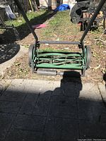 Green Lee Valley mechanical shearing lawnmower shown front-facing, positioned on paved ground with garden and yard background.