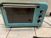 Front view of teal classic toaster oven showing glass door, silver handle, and set of three control knobs on the right side.
