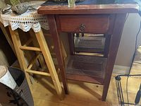 Brown wooden side table with drawer and decorative metal mesh shelf insert, placed on a wooden floor next to a light wooden stool.