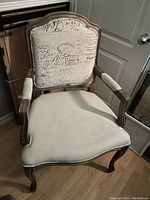 Photo of an upholstered armchair showing the script-style fabric on the backrest and plain light fabric on seat and armrests, wooden frame with carved legs.