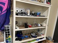 White shelving unit with assorted kitchenware items including glass bowls, teapots, paper towel rolls, and a black hot plate.