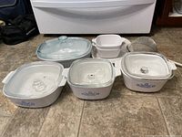 Photo of all bakeware pieces including covered blue cornflower and French white baking dishes, nested white square dishes, and metal strainer on floor.