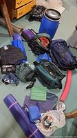 Overall view of camping gear assortment including knapsacks, storage bin, mats and accessories on floor