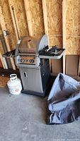 Full view of the Coleman propane BBQ in a garage, showing the grill with side racks, propane tank beneath, and a fabric cover on the floor beside it.