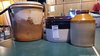 Photo of three vintage earthenware crocks on a table. Left crock is 5-gallon sized with a lid (lid cracked), middle is a medium oval crock with dark brown rim, right is smaller with two-tone brown and beige color and narrow opening.