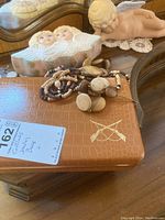 View of wooden bead necklaces, cufflinks, earrings on a brown faux leather jewelry box with angel-themed jewelry box and porcelain angel figure in background.