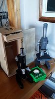 Photo of two microscopes side by side on a table; one black Bushnell with adjustable knobs and a wooden case behind, and one gray Lumex microscope with a tall viewing tube