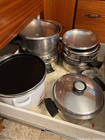 Drawer containing the collection of pots and pans including large stainless steel pots with lids, multiple stacked stainless steel pans with black handles, and a white enamel pot with red stripes.