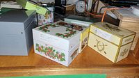 Three decorative metal and cardboard recipe card boxes with labels and kitchen items in background