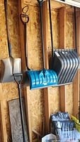 Five snow removal tools shown hanging on a wooden wall, including three snow shovels with D-handles, one large blue plastic snow scoop, and one straight edge shovel with a flat blade.