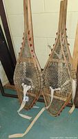 Two pairs of vintage wooden snowshoes leaning against a wall, showing rawhide webbing and cloth straps