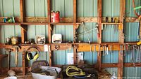 Wide view of tools and hardware on wooden shelves and hooks against a metal wall, showing clamps, wrenches, sockets, and other hand tools.
