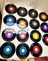 Image showing 17 loose 45 RPM vinyl records with various colored labels from studios like RCA Victor, Columbia, Capitol, MGM, Decca, Coral, including several colors such as red, blue, yellow, orange, purple.