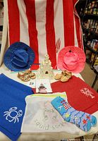 Photo shows a large red and white striped Beach Resort Cabana towel, two children's safari hats (blue with dolphin brim and pink with hibiscus brim), two pairs of children's sandals, blue and red placemats with crab and lobster logos, squid socks in mesh bag, and a handled lighthouse bag.