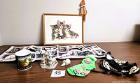 Photo showing the full lot with the embroidered kittens framed picture, tapestry runner, Lenox salt and pepper set with tray, soapstone mouse, cat face mask, tabby cat mug, wooden cat figure, and cat socks laid out on table.