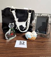 Four cat-themed items displayed on wooden surface: hand-blown glass cat paperweight with goldfish, ceramic Siamese kitten figurine in a basket, metal picture frame labeled 'Meow,' black purse with white cat designs.
