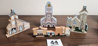 Four Cats Meow wooden miniature churches displayed on table: Church of Saint Ann, Sacred Heart Church, Holy Trinity R.C. Church, and Holy Family School of St Ann's Church. Each shows detailed painted architecture typical of Cats Meow collectibles.